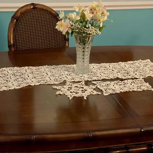May include: A wooden table with a white crocheted table runner and two smaller crocheted placemats. The table is set with a vase of white flowers.