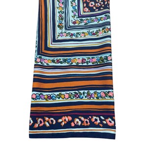 May include: Folded scarf with a colorful floral and striped pattern. The scarf features a square design with a navy blue background, orange and white stripes, and floral accents in pink, yellow, and blue.