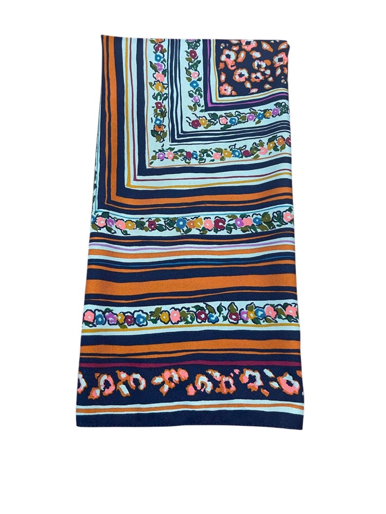 May include: Folded scarf with a colorful floral and striped pattern. The scarf features a square design with a navy blue background, orange and white stripes, and floral accents in pink, yellow, and blue.