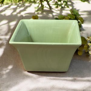 May include: A square, light green ceramic bowl with a slightly flared design. The bowl has a smooth, glossy finish and is empty. The bowl is sitting on a neutral-colored surface, with greenery in the background.