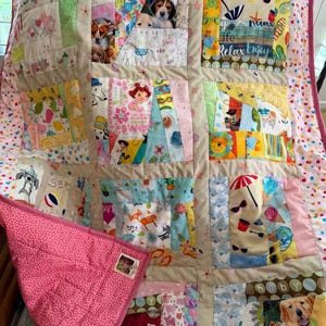 May include: A patchwork quilt featuring a variety of colorful fabric squares with prints of animals, beach scenes, and cartoon characters. The quilt is bordered by a pink fabric with small dots and has a solid pink backing with a small square label.