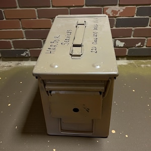 May include: An olive drab metal ammunition box with a hinged lid and a handle. The box has the words "Harbor Services" and "220 Cordless Drill Rest" written on it in black marker. The box is rectangular with a latch on the front.