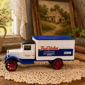 May include: A die-cast metal toy delivery truck featuring a white cab and box body with blue accents and red wheels. The side of the truck displays the text True Value Hardware 10th Anniversary Edition and Your Store of First Choice.