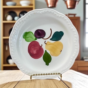 May include: White ceramic serving dish with a scalloped edge and a hand-painted fruit design. The fruit includes a red apple, yellow pear, and purple plum with green leaves. The dish is displayed on a gold-colored metal stand.