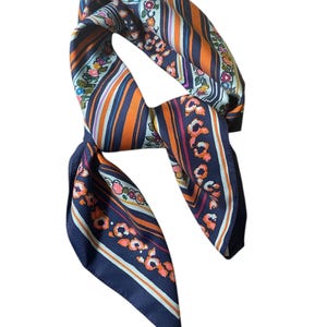 May include: A silk scarf with a floral and striped pattern. The scarf features a navy blue background with orange, white, and pink stripes and floral accents. The scarf is folded and tied, showcasing its design.