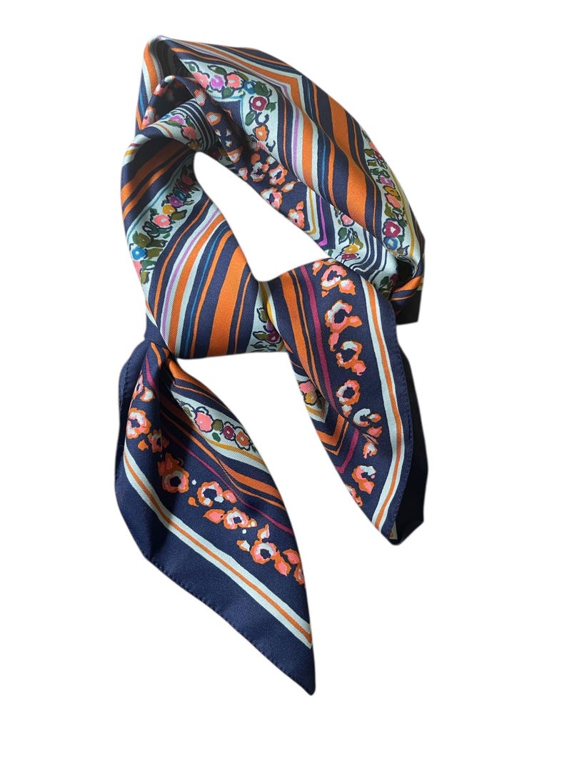 May include: A silk scarf with a floral and striped pattern. The scarf features a navy blue background with orange, white, and pink stripes and floral accents. The scarf is folded and tied, showcasing its design.