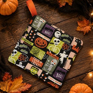 May include: A square fabric pot holder featuring a black background with a colorful Halloween pattern including skeletons, mummies, pumpkins, bats, ghosts, and black cats. The text reads BOO!, Jack-o-Lantern, Frankenstein's Monster, The Mummy, and TRICK OR TREAT? with an orange hanging loop.