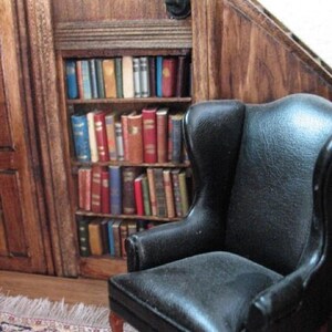 May include: Miniature library scene with a black leather wingback chair and a bookshelf filled with tiny books. The bookshelf is built into a wooden wall, and a small rug is on the floor. A wall-mounted candle holder is above the bookshelf.