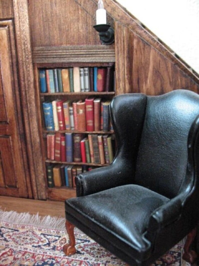 May include: Miniature library scene with a black leather wingback chair and a bookshelf filled with tiny books. The bookshelf is built into a wooden wall, and a small rug is on the floor. A wall-mounted candle holder is above the bookshelf.