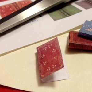 May include: A collection of miniature books, including a red book with a patterned cover and a blue book with gold floral designs. A pair of scissors and a sheet of paper with book cover designs are also visible.