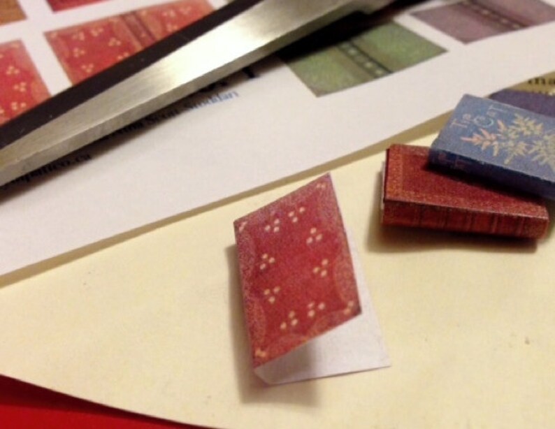 May include: A collection of miniature books, including a red book with a patterned cover and a blue book with gold floral designs. A pair of scissors and a sheet of paper with book cover designs are also visible.