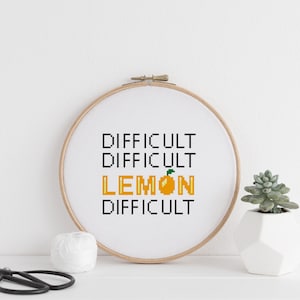 May include: A cross-stitch embroidery hoop featuring the text "DIFFICULT DIFFICULT LEMON DIFFICULT" in black and yellow pixelated font, with a yellow pixelated lemon graphic. The hoop is displayed against a white backdrop.