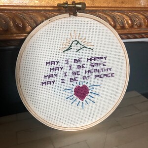 May include: A round, cross-stitched embroidery hoop with the words "MAY I BE HAPPY, SAFE, HEALTHY, AT PEACE." The design includes a mountain with a rising sun, a heart, and radiating lines, all in various colors.