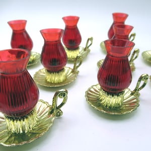 May include: Several red glass candle holders with a ribbed design. Each holder is set on a gold-coloured, leaf-shaped base with a decorative handle. The candle holders are arranged on a white surface.