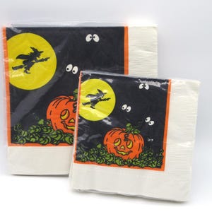May include: Two packs of Halloween-themed paper napkins. Each napkin features a black background with a yellow moon, a witch on a broom, an orange jack-o'-lantern, and green foliage. White napkins are visible.