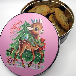 Puede incluir: Una lata de metal rosa con un tema navideño que presenta un ciervo, un árbol de Navidad y las palabras "Christmas Joys". La lata está abierta y llena de galletas con chispas de chocolate.