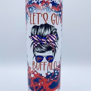 May include: A white tumbler with a red, white, and blue splatter design. The tumbler features a graphic of a woman with a messy bun, a red, white, and blue bandana, and sunglasses that say "Bills" on the lenses. The text "Let's Go Buffalo" is printed on the tumbler.
