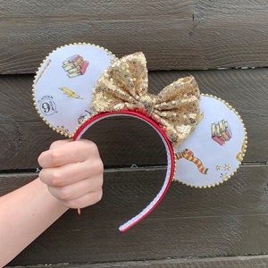 May include: A pair of white mouse ears with a red headband. The ears are decorated with gold glitter, a gold bow, and a print of books, stars, and the text "Platform 9 3/4".