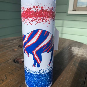 May include: A white stainless steel tumbler with a red and blue splatter design. The tumbler features a blue and white buffalo logo. A clear straw is inserted into the lid.