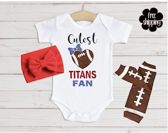 May include: A white baby bodysuit with the text "Cutest Titans Fan" and a football-shaped heart. Includes a red bow headband and brown and white football-themed leg warmers. The bodysuit is hanging on a wooden hanger.