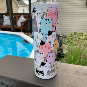 May include: A white stainless steel tumbler with a colorful cat print. The tumbler has a black lid and a straw. The cats are in various colors, including pink, blue, yellow, and green.
