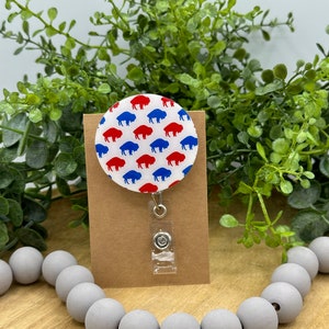 May include: A white round badge holder with a red and blue buffalo pattern. The badge holder is attached to a clear retractable badge reel with a silver clip.