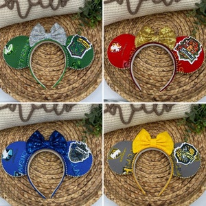 May include: Four pairs of Harry Potter themed Minnie Mouse ears. Each pair features a different Hogwarts house crest: Gryffindor, Slytherin, Hufflepuff, and Ravenclaw. The ears are decorated with glitter and bows.