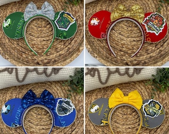 May include: Four pairs of Harry Potter themed Minnie Mouse ears. Each pair features a different Hogwarts house crest: Gryffindor, Slytherin, Hufflepuff, and Ravenclaw. The ears are decorated with glitter and bows.