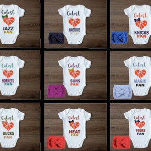 May include: Nine white baby onesies with a basketball heart graphic and the text "Cutest" above the heart. Each onesie has a different NBA team name below the heart. The onesies are paired with a matching headband in a variety of colors.