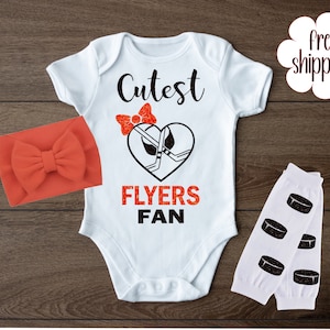 K&ouml;nnte beinhalten: Wei&szlig;er Baby-Strampler mit dem Text "Cutest Flyers Fan" und einer roten Schleife mit einem Hockeystock- und Puck-Design. Der Strampler wird mit einem roten Stirnband und wei&szlig;en Beinstulpen mit schwarzen Hockey-Puck-Designs kombiniert.
