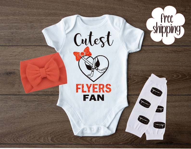 K&ouml;nnte beinhalten: Wei&szlig;er Baby-Strampler mit dem Text "Cutest Flyers Fan" und einer roten Schleife mit einem Hockeystock- und Puck-Design. Der Strampler wird mit einem roten Stirnband und wei&szlig;en Beinstulpen mit schwarzen Hockey-Puck-Designs kombiniert.