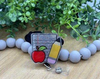 May include: A silver glitter badge holder with a black lanyard. The badge holder features a cartoon image of a red apple, a yellow pencil, and a sheet of lined paper.