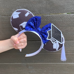 May include: Black and white graduation cap themed Minnie Mouse ears with a blue bow and a white tassel. The cap has the year 2020 printed on it.
