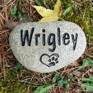 May include: A gray, oval-shaped memorial stone with the name "Wrigley" carved in black, along with a heart and paw print design. The stone rests on a bed of brown and green foliage.