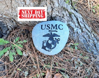 May include: A smooth, light gray river stone featuring the black engraved text USMC above the official United States Marine Corps eagle, globe, and anchor emblem. The stone has a rounded, irregular shape and a matte finish with clear, dark markings.