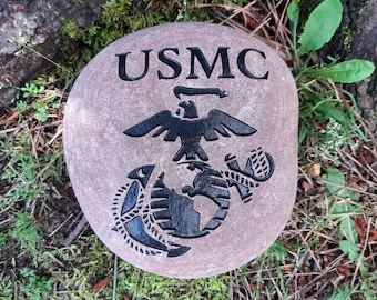 May include: A round, tan stone with the letters "USMC" and the United States Marine Corps emblem carved in black. The emblem features an eagle, globe, and anchor. The stone is surrounded by green grass and moss.