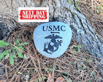 May include: A smooth, light grey river stone featuring the black engraved text USMC above the United States Marine Corps eagle, globe, and anchor emblem. A white rectangular sign with red text reading NEXT DAY SHIPPING is positioned above.