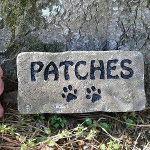 May include: A gray concrete stone with the word "PATCHES" painted in black letters. Two black paw prints are painted below the name.