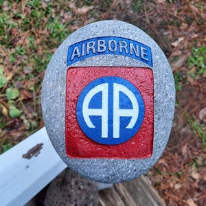 May include: A gray stone painted with the 82nd Airborne Division insignia. The insignia is a red square with a blue circle containing the letters "AA" in white. The words "AIRBORNE" are painted in blue on a blue banner above the insignia.