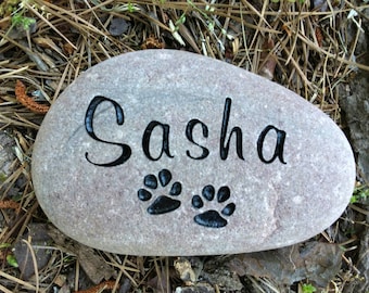 May include: A gray stone with the name "Sasha" written in black cursive lettering and two black paw prints.