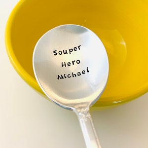 May include: A silver soup spoon with the words "Souper Hero Michael" engraved on the front. The spoon is resting in a yellow bowl.