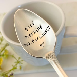 May include: A silver spoon with the words "good morning ms. fernandez" and a heart symbol stamped on the bowl. The spoon is resting in a white mug. The spoon has a decorative handle.