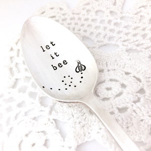 May include: A silver spoon with the words "let it bee" stamped on the bowl, along with a small bee illustration and dotted trail. The spoon rests on a white, lacy surface.