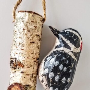 May include: A wooden bird ornament, painted black and white with white spots, is hanging from a piece of twine. The bird is perched on a small, wooden birch log ornament, also hanging from twine.