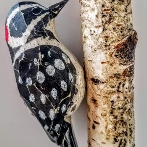 May include: A wooden bird ornament with black and white markings and a red beak, hanging from a birch bark log ornament.