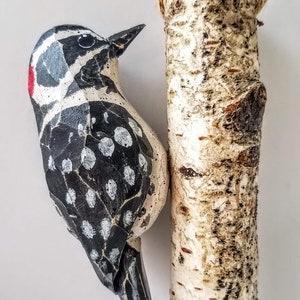 May include: A wooden carved woodpecker with black and white markings and a red spot on its head, hanging from a birch log with a twine string.