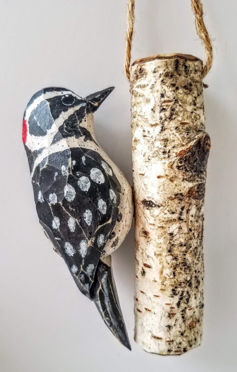 May include: A wooden carved woodpecker with black and white markings and a red spot on its head, hanging from a birch log with a twine string.