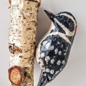 May include: A wooden bird ornament with black and white paint and a red beak, hanging from a birch log ornament with a rope loop.