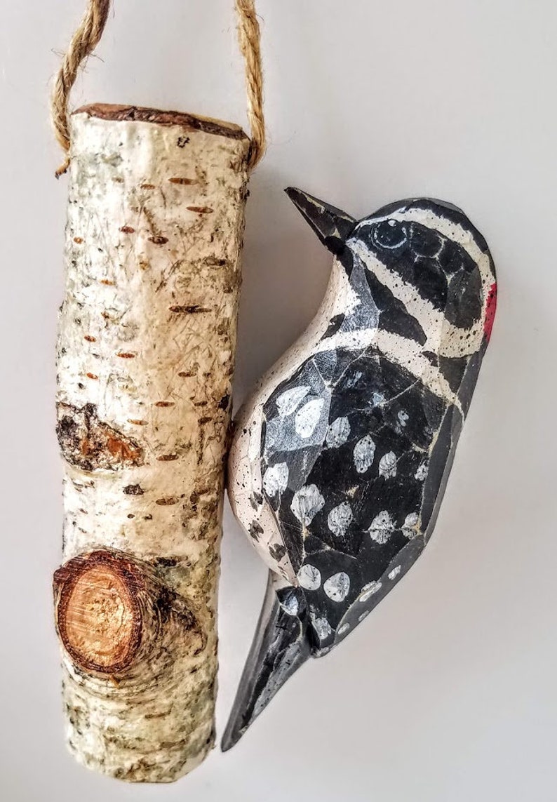 May include: A wooden bird ornament with black and white paint and a red beak, hanging from a birch log ornament with a rope loop.