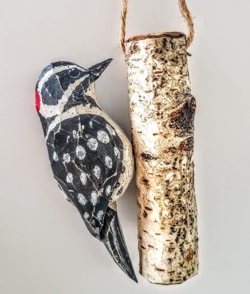 May include: A hand-painted wooden bird ornament with black and white markings and a red beak, hanging from a birch branch.